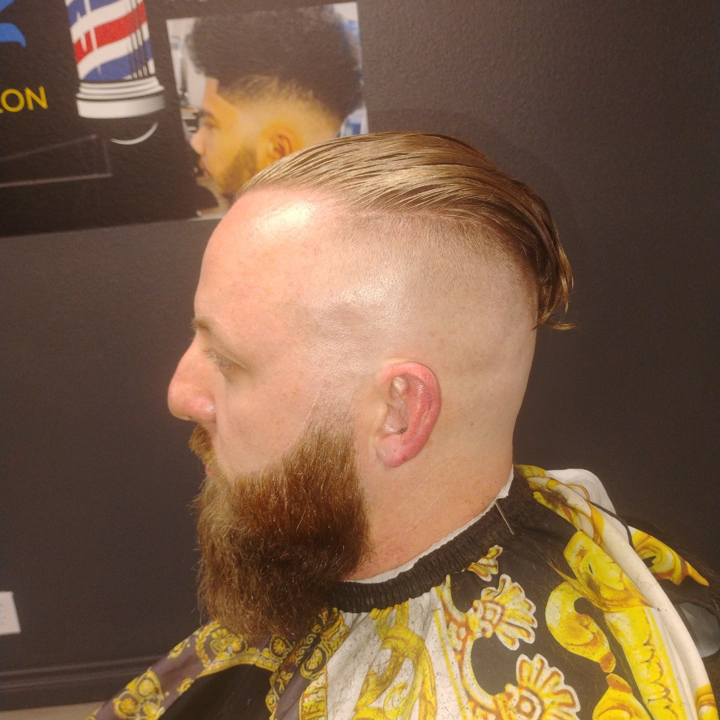 Slick back with high skin fade, full beard line-up, gold-pattern cape.